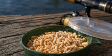 Frische Angelmaden in einer Köderdose neben einer Angelrute am Wasser