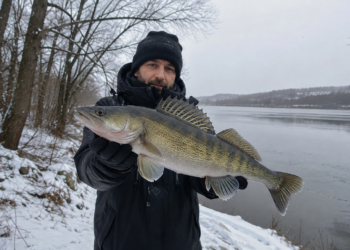 Angler hält einen großen Zander an einem verschneiten Gewässer in die Kamera.