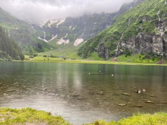 Bergsee bei Regen unter tief hängenden Wolken und grünen Berghängen