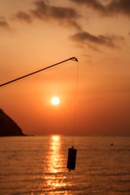 Zylindrisches Angelgewicht an einer Schnur unterhalb der Rutenspitze vor einem orangefarbenen Sonnenuntergang am Meer.