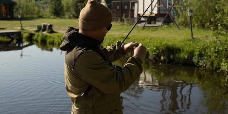 Angler mit Mütze und Sonnenbrille bereitet seine Angelmontage am Ufer eines Teichs vor.