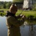 Angler mit Mütze und Sonnenbrille bereitet seine Angelmontage am Ufer eines Teichs vor.
