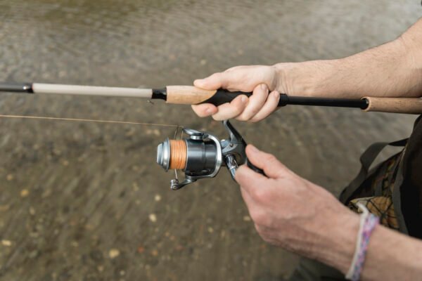 Hände eines Anglers an einer Spinnrute mit grauer Stationärrolle und orangefarbener Schnur über flachem Wasser.