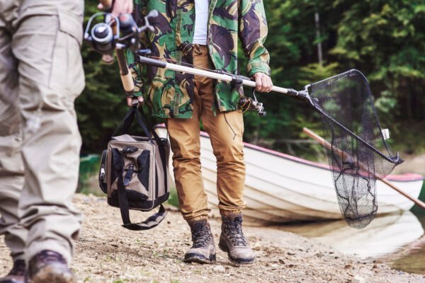 Angler mit kompakter Angeltasche, Angelrute und Kescher am Ufer eines Sees.