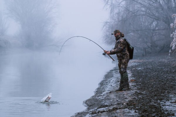 Angler drillt im Thermoanzug einen Fisch am nebligen und verschneiten Flussufer.