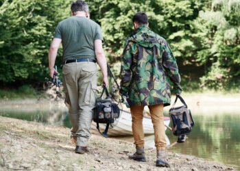 Zwei Angler tragen Angeltaschen, Kescher und Angelruten zu einem Boot am See.