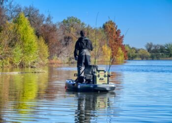 Angler auf einem Angelkajak mit mehreren aufrecht in Rutenhaltern verstauten Angelruten vor einem herbstlichen Ufer.