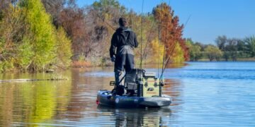 Angler auf einem Angelkajak mit mehreren aufrecht in Rutenhaltern verstauten Angelruten vor einem herbstlichen Ufer.