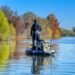 Angler auf einem Angelkajak mit mehreren aufrecht in Rutenhaltern verstauten Angelruten vor einem herbstlichen Ufer.