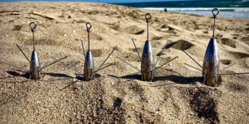 Vier längliche Brandungsbleie mit Drahtkrallen und Befestigungsösen stehen nebeneinander im Strandsand, dahinter ist das Meer sichtbar.