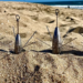 Vier längliche Brandungsbleie mit Drahtkrallen und Befestigungsösen stehen nebeneinander im Strandsand, dahinter ist das Meer sichtbar.