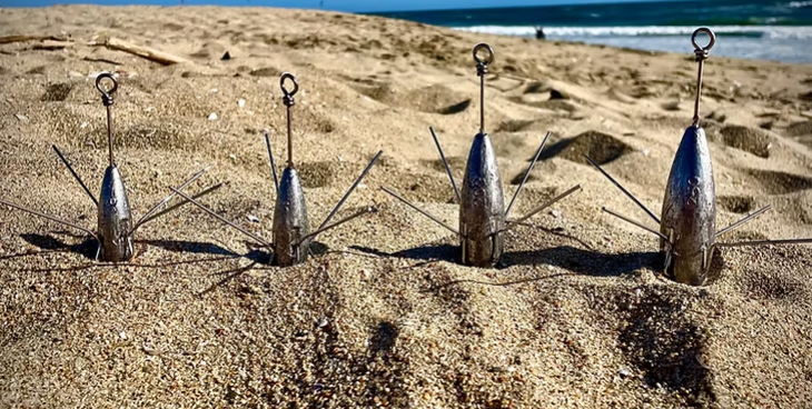 Vier längliche Brandungsbleie mit Drahtkrallen und Befestigungsösen stehen nebeneinander im Strandsand, dahinter ist das Meer sichtbar.