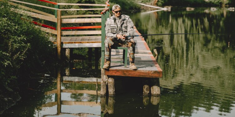 Angler sitzt mit einer Angelrute auf einem Holzsteg an einem ruhigen See.