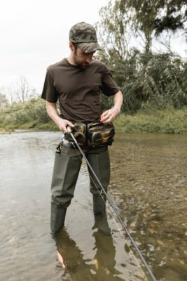 Angler mit Angelrute und geöffneter Hüfttasche beim Fischen in einem flachen Fluss
