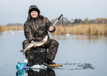 Angler im Thermoanzug sitzt beim Eisangeln mit gefangenem Hecht auf einem zugefrorenen See.
