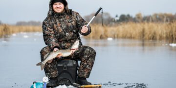 Angler im Thermoanzug sitzt beim Eisangeln mit gefangenem Hecht auf einem zugefrorenen See.