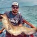 Angler hält einen großen, selbst gefangenen Wels mit beiden Händen auf einem Boot.