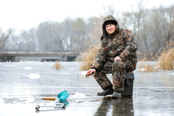 Angler in einem gefütterten Tarnanzug sitzt beim Eisangeln neben einem gebohrten Eisloch.
